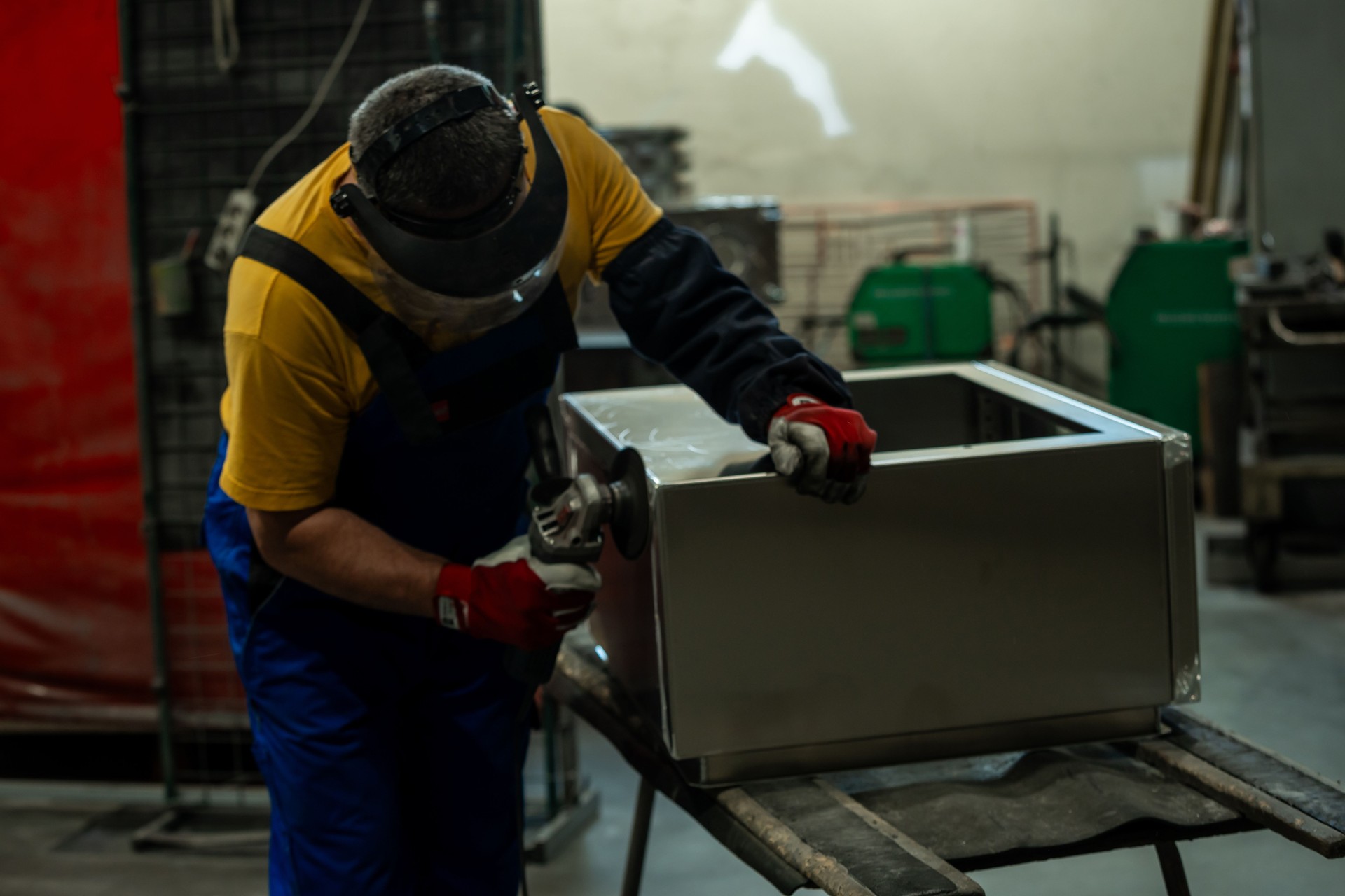 A focused worker wearing a face shield, gloves, and overalls uses a handheld power tool to grind a metal box on a workbench in a well lit factory.